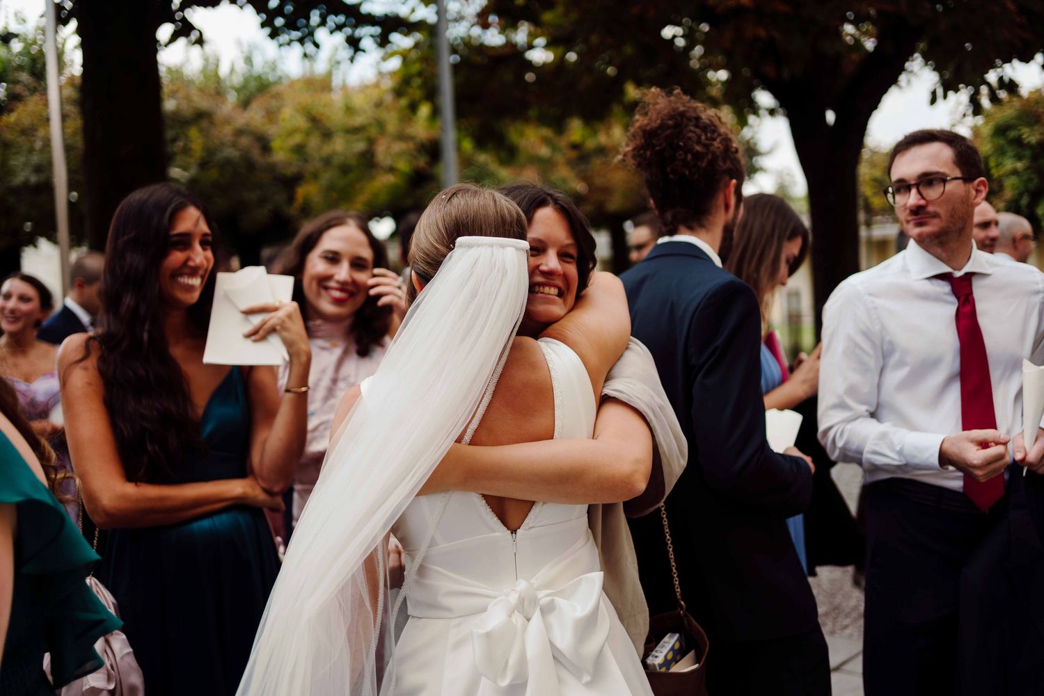 Bride in white gown hugging guest at outdoor wedding reception with smiling attendees in background.