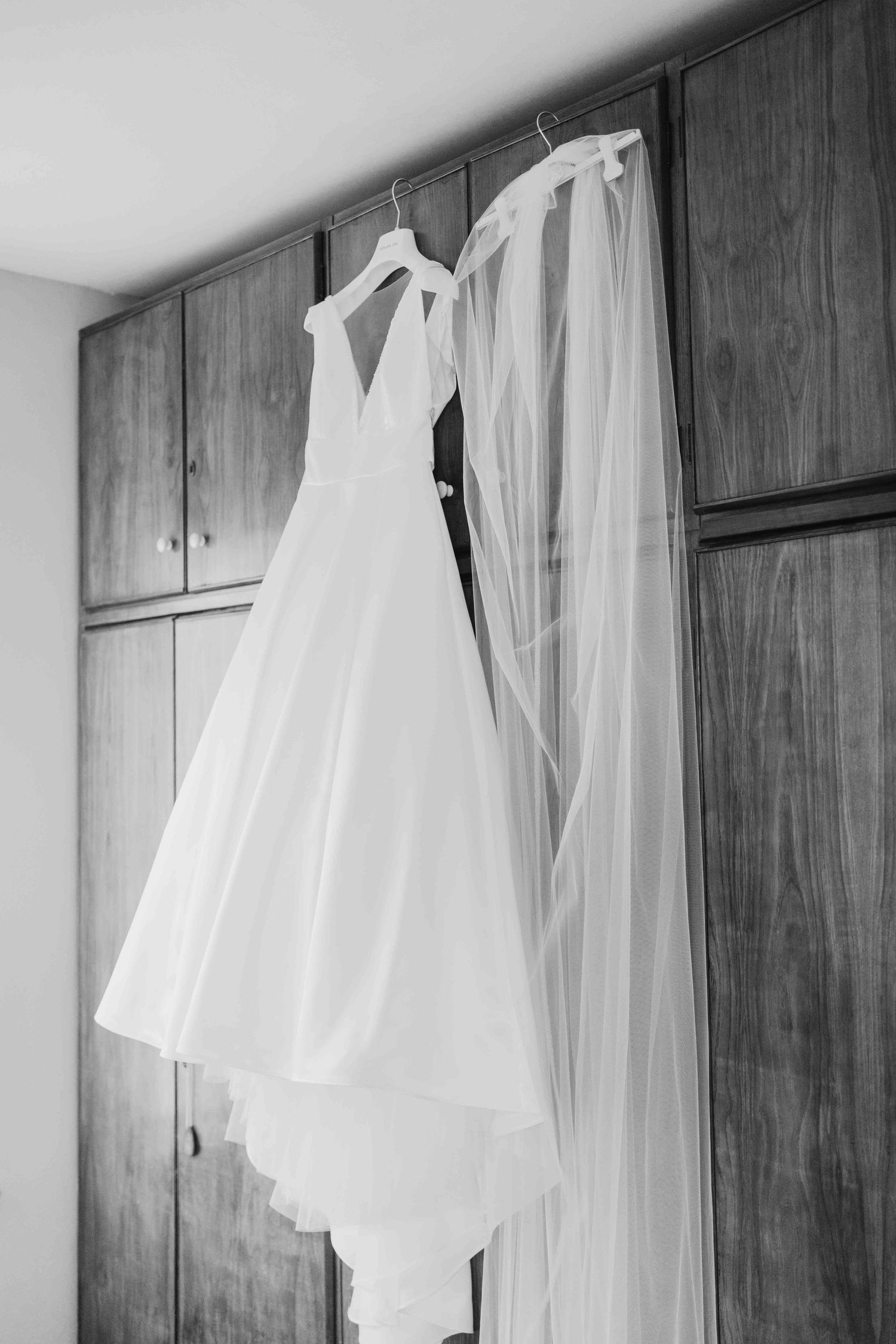 Black and white photo of a wedding dress and veil hanging on wooden wardrobe doors.