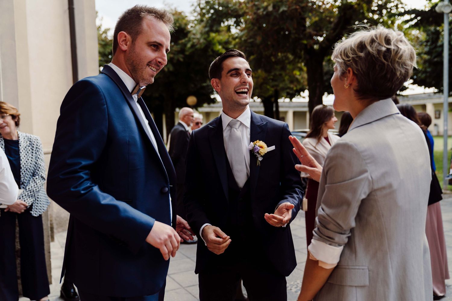 Two men in suits laugh with a woman at an outdoor wedding reception celebration.