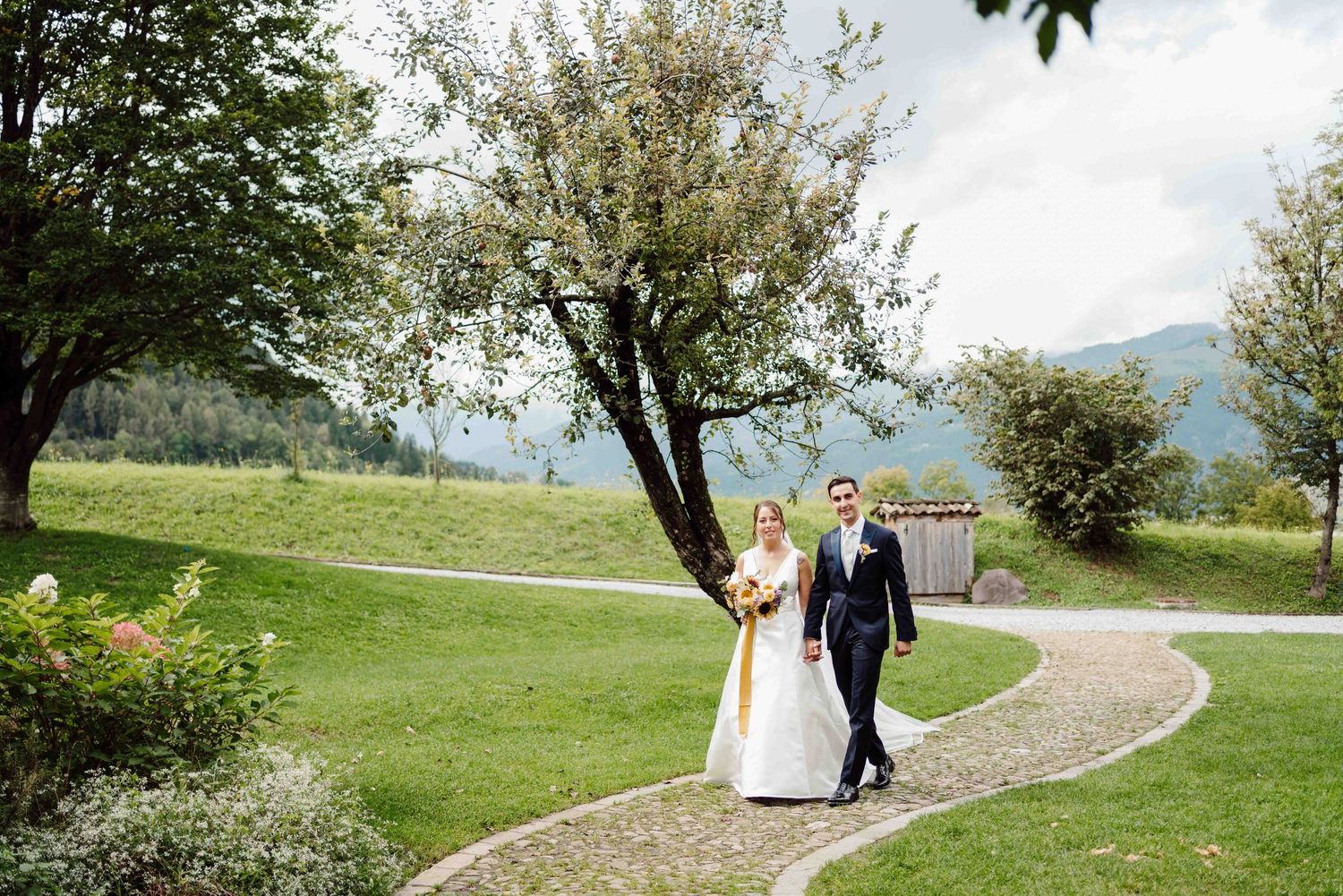 A bride and groom walk hand in hand along a winding path through lush green gardens with mountain views.