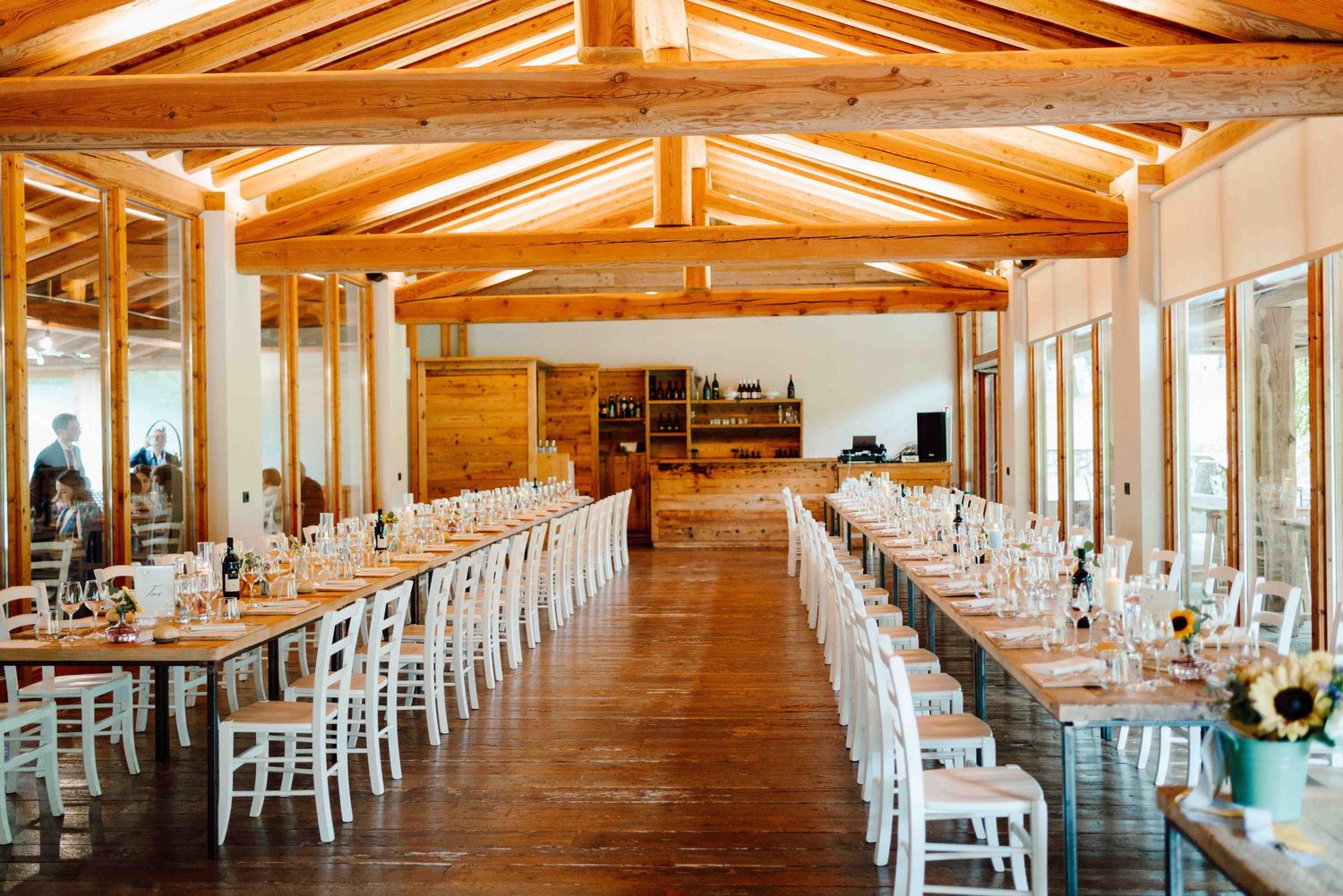 Rustic wedding reception venue with long banquet tables, white chairs, wooden beamed ceiling, and elegant table settings.