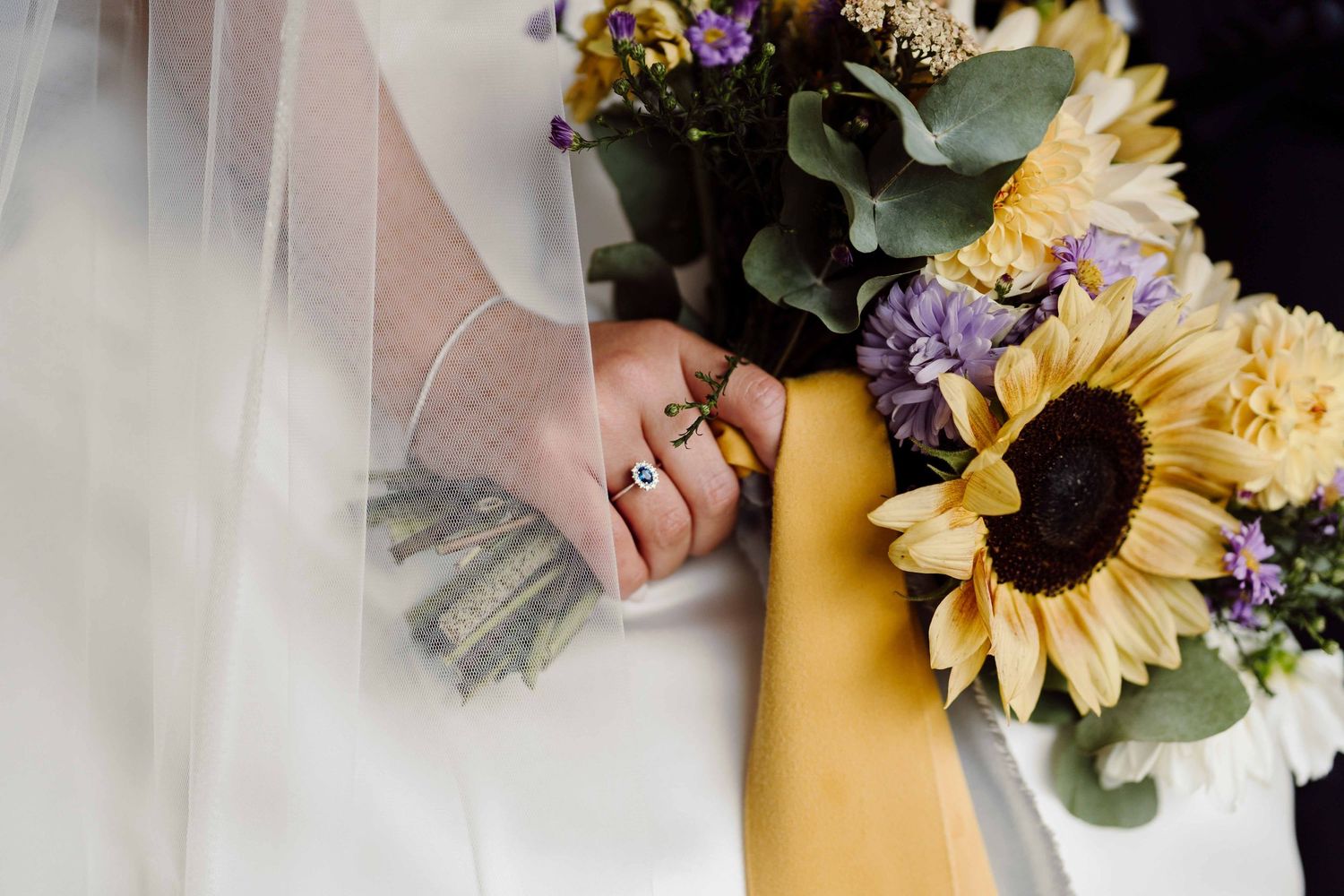 Bride holding a sunflower bouquet with purple flowers, wearing gold rings, draped in a white wedding veil.