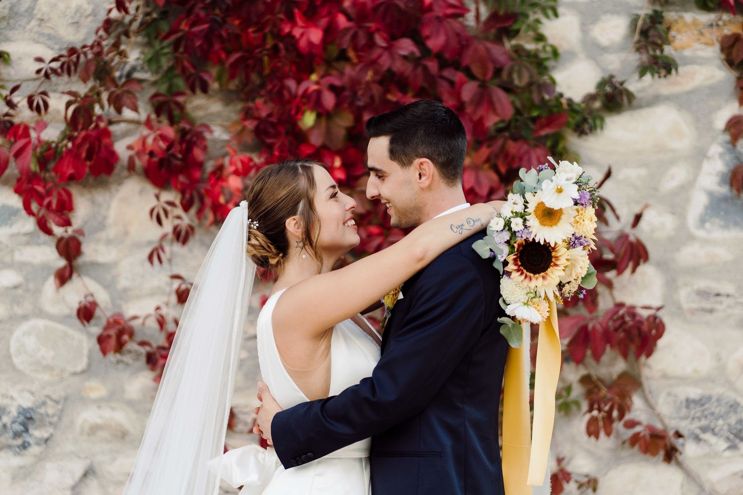 Bride and groom embrace before red autumn ivy on stone wall, bride holding sunflower bouquet with yellow ribbon.