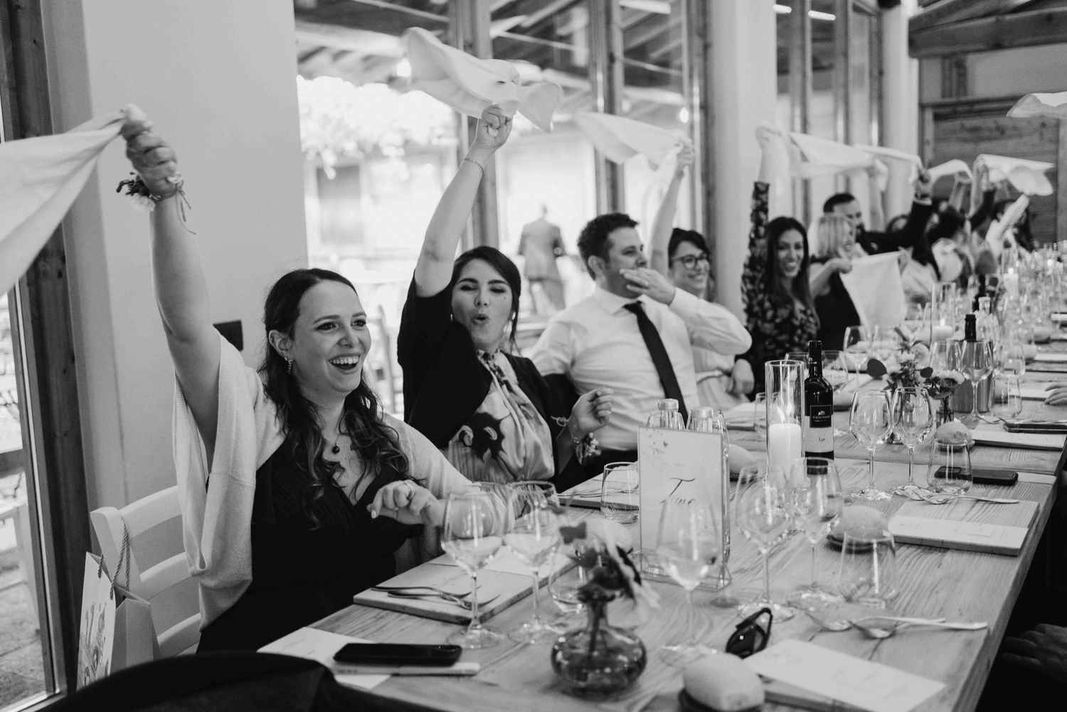 Black and white photo of wedding guests laughing and raising napkins at a long reception dinner table.