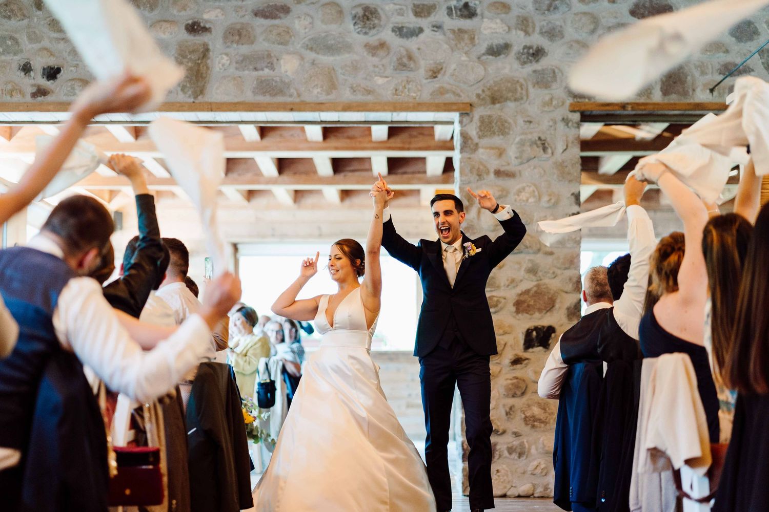 Bride and groom celebrate their wedding reception entrance with guests waving white ribbons in a rustic stone venue.