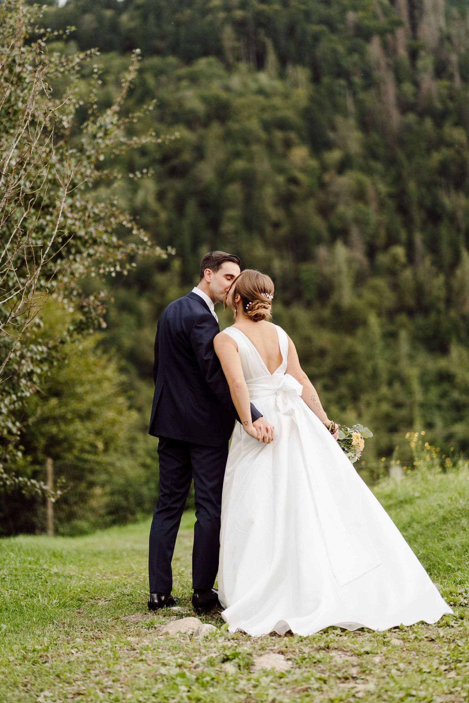 Bride and groom share a tender kiss outdoors, surrounded by lush green forest, bride in elegant white gown holding bouquet.