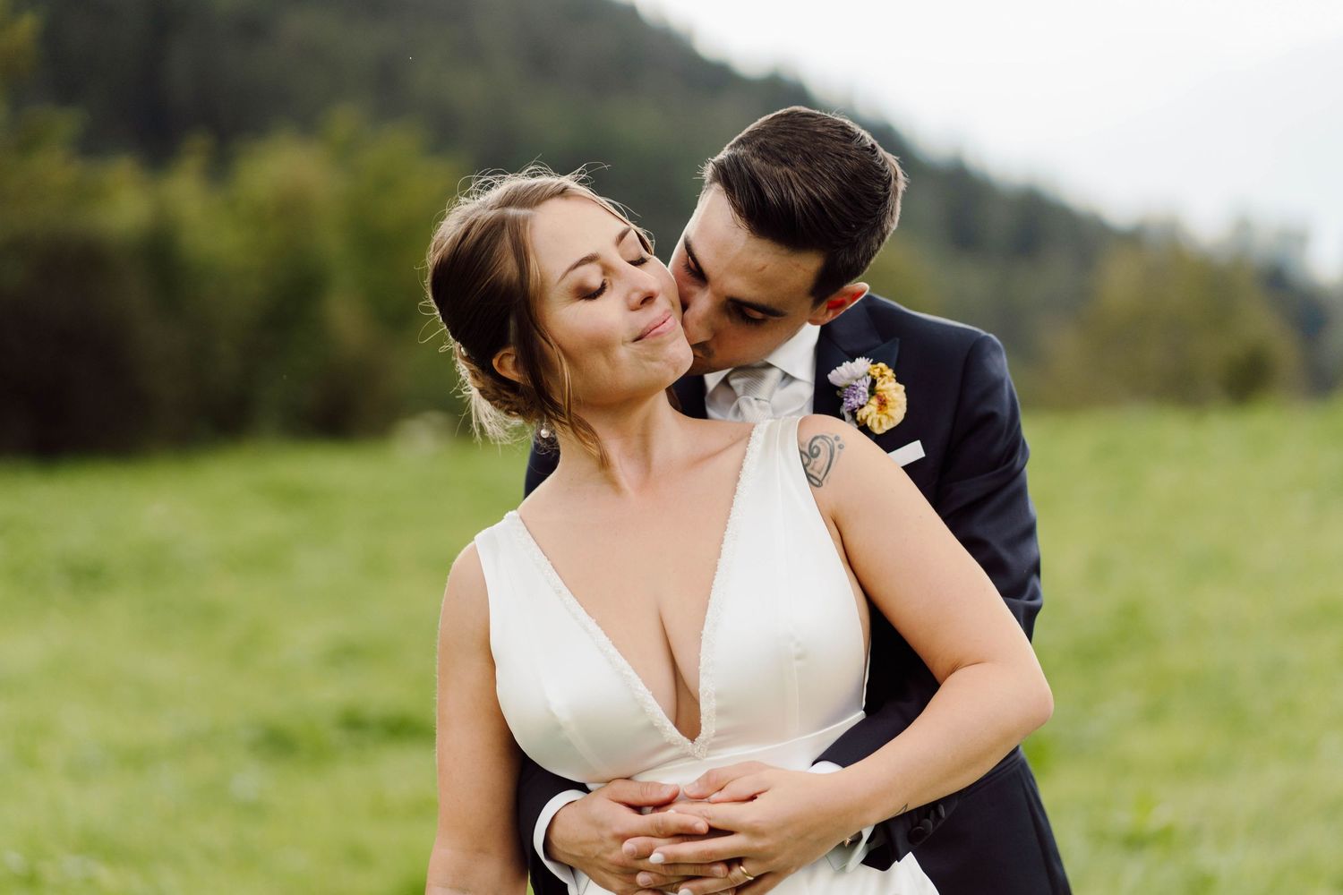 Bride and groom share a romantic embrace in a lush green meadow, groom kissing bride's cheek in wedding attire.