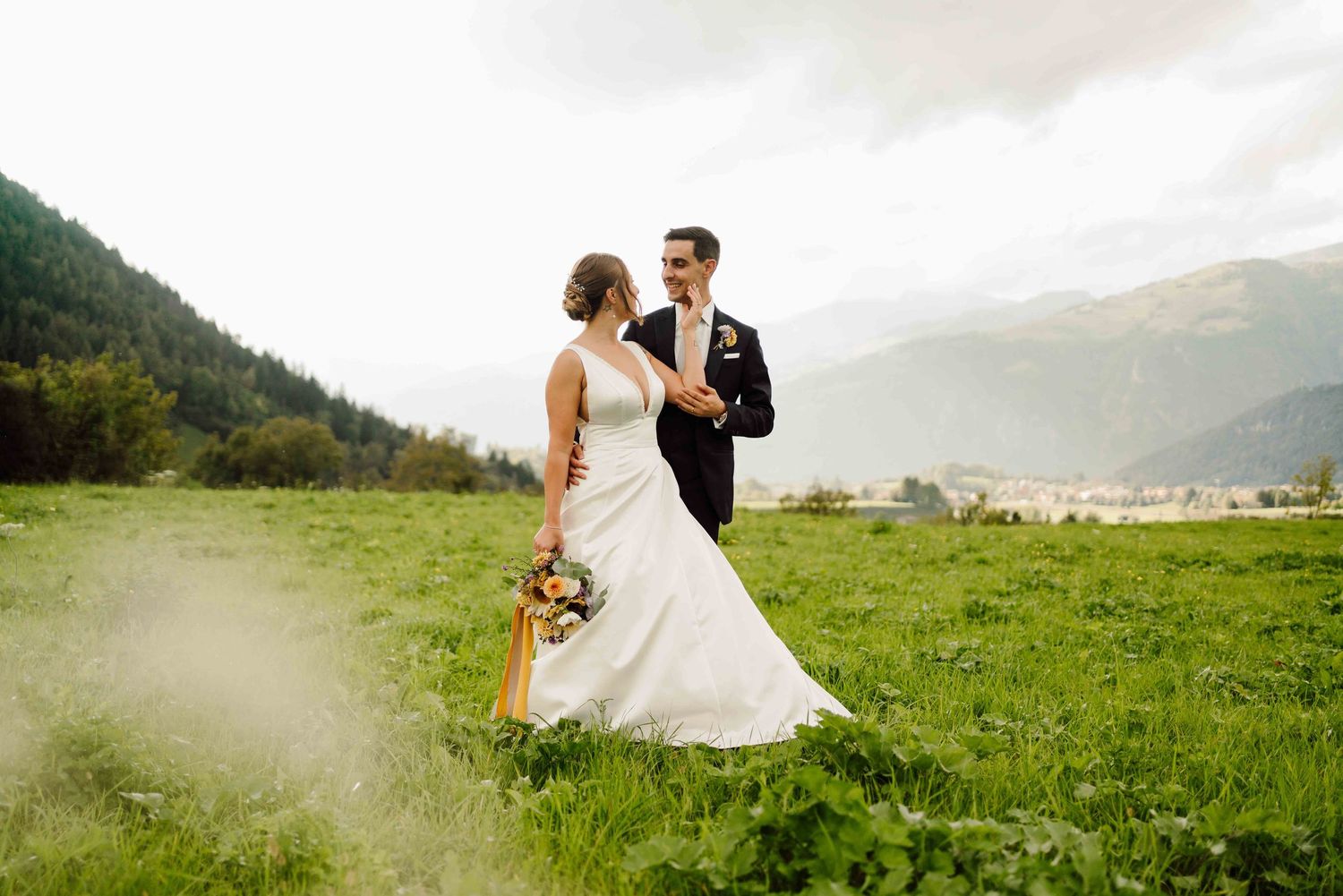 Bride and groom share a romantic moment on a lush green meadow with stunning mountain backdrop on their wedding day.