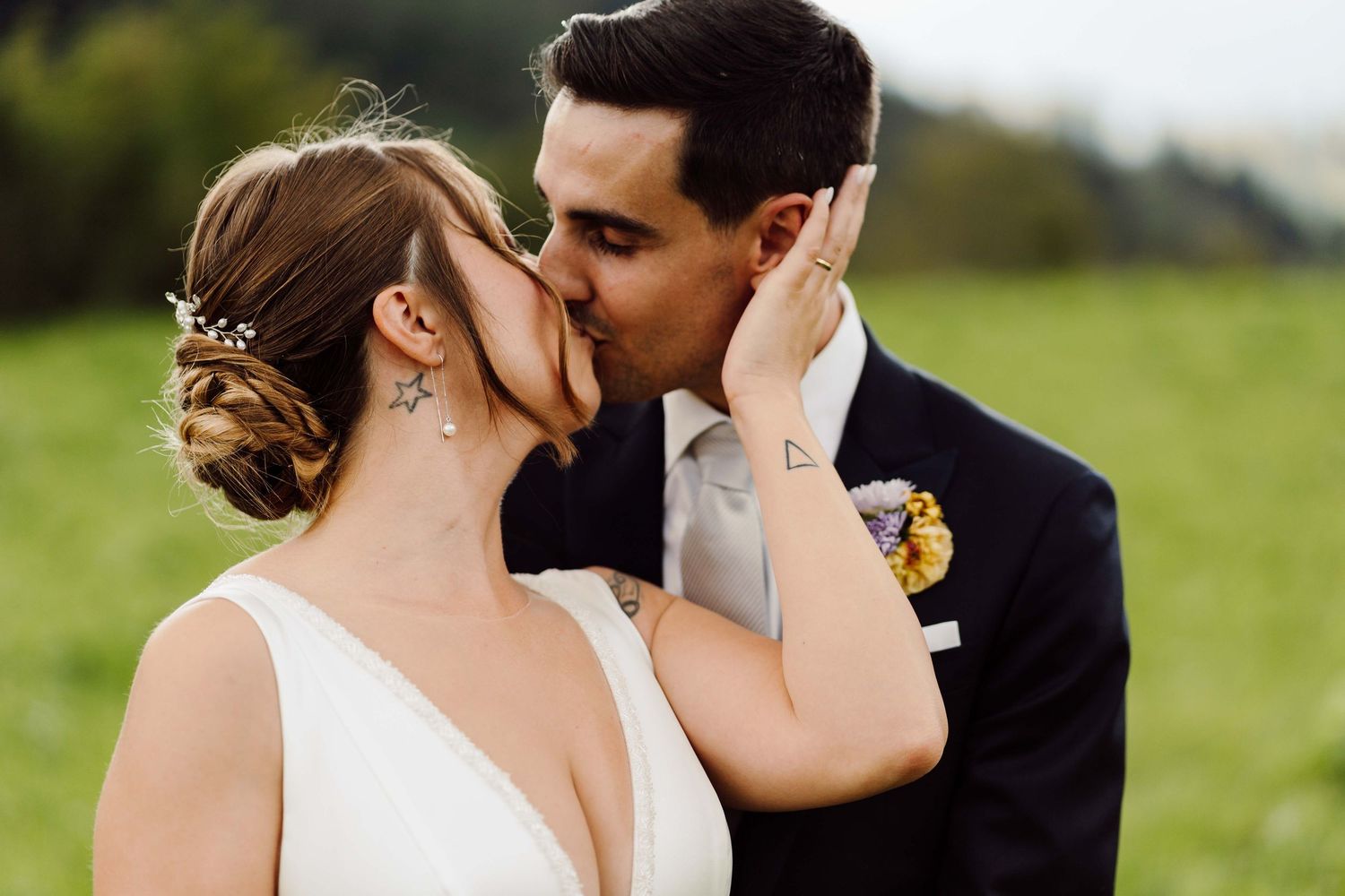 Bride and groom sharing a romantic kiss outdoors on their wedding day, with green fields in the background.