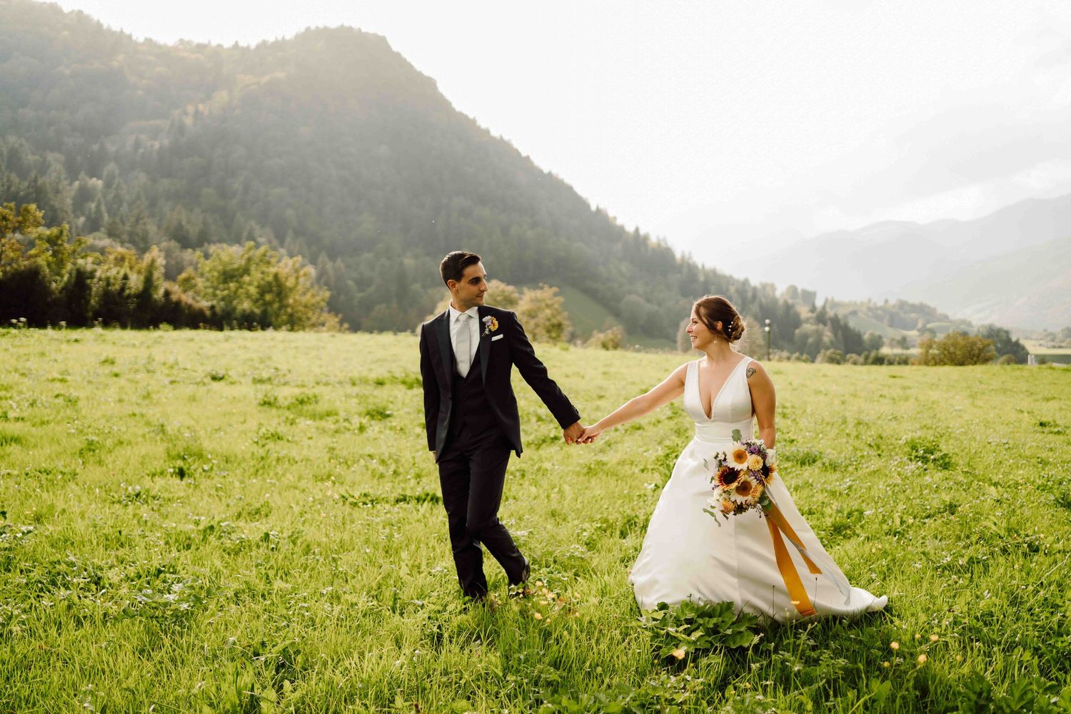 Bride and groom walking hand in hand through a lush green meadow with mountain backdrop on their wedding day.