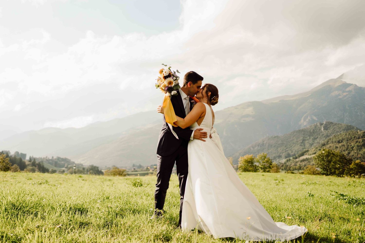 Bride and groom kissing in a green meadow with stunning mountain backdrop on their wedding day.