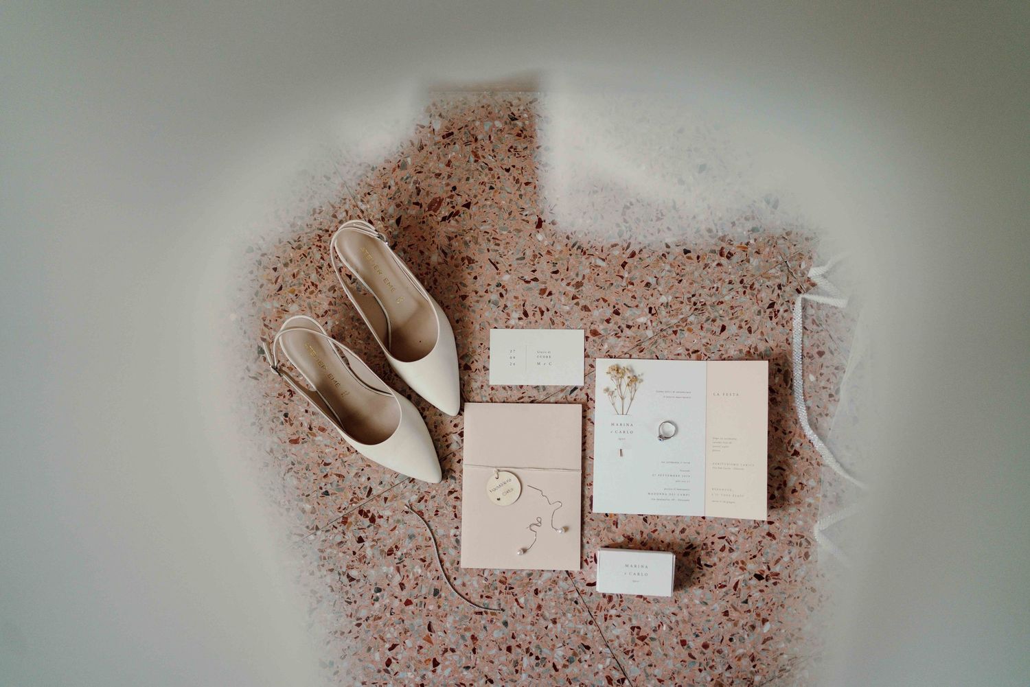 Elegant wedding flat lay featuring blush invitation suite, rings, and bridal shoes on terrazzo surface.