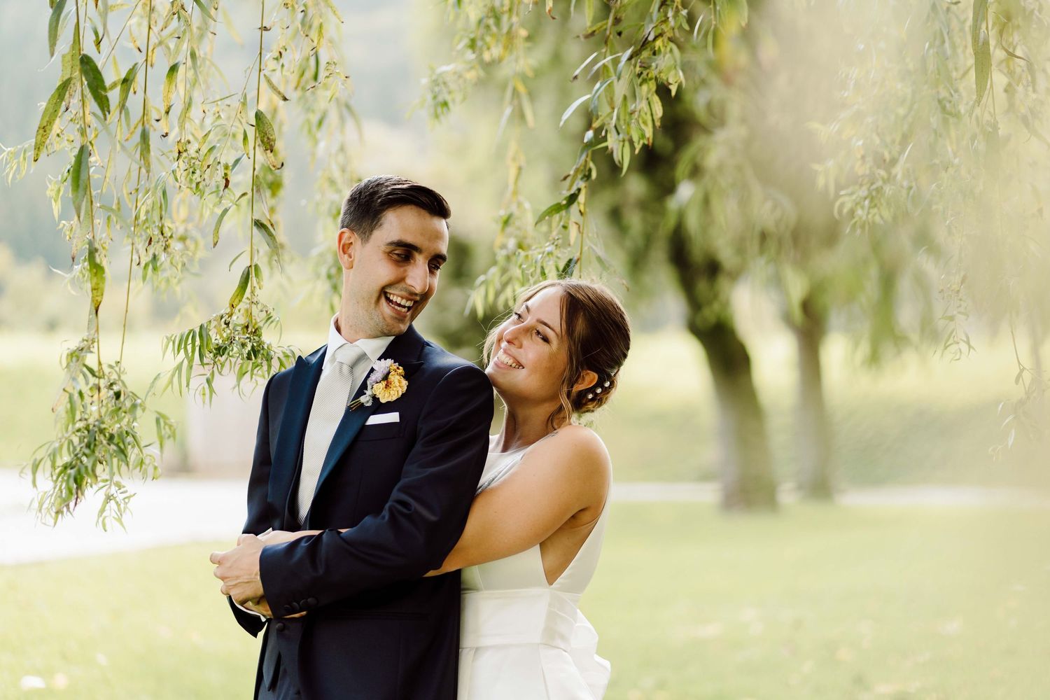 A bride and groom embrace under a weeping willow tree, smiling joyfully on their wedding day outdoors.