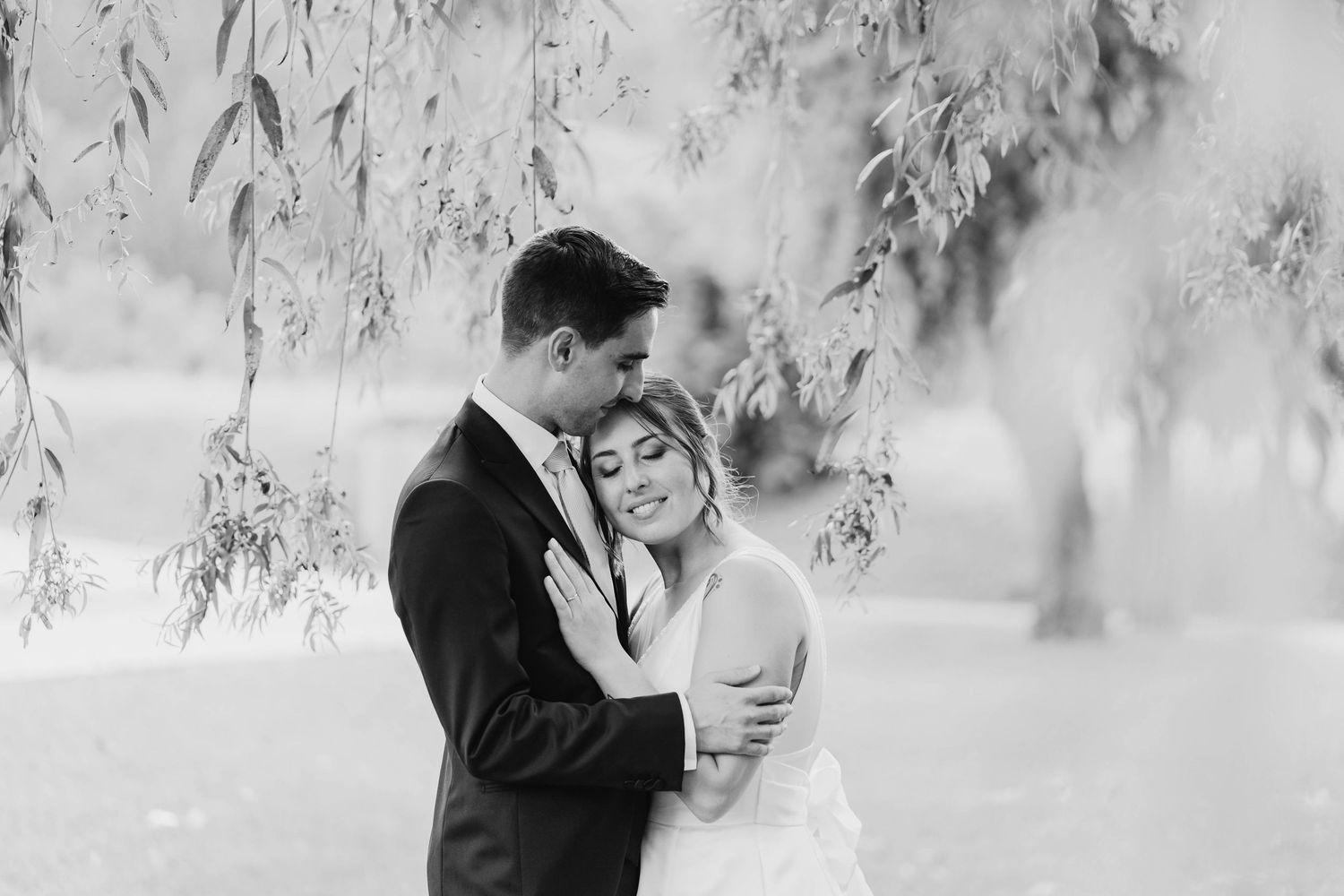 Black and white photo of a bride and groom embracing tenderly under weeping willow branches.
