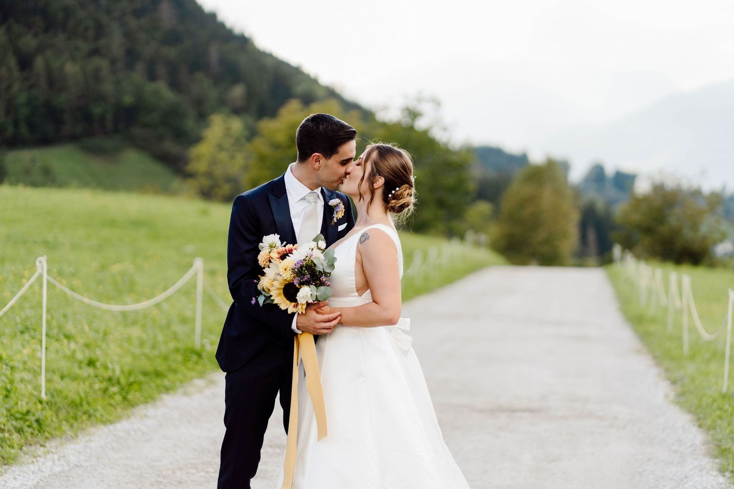 Bride and groom share a romantic kiss on a scenic path surrounded by lush green meadows and mountains on their wedding day.
