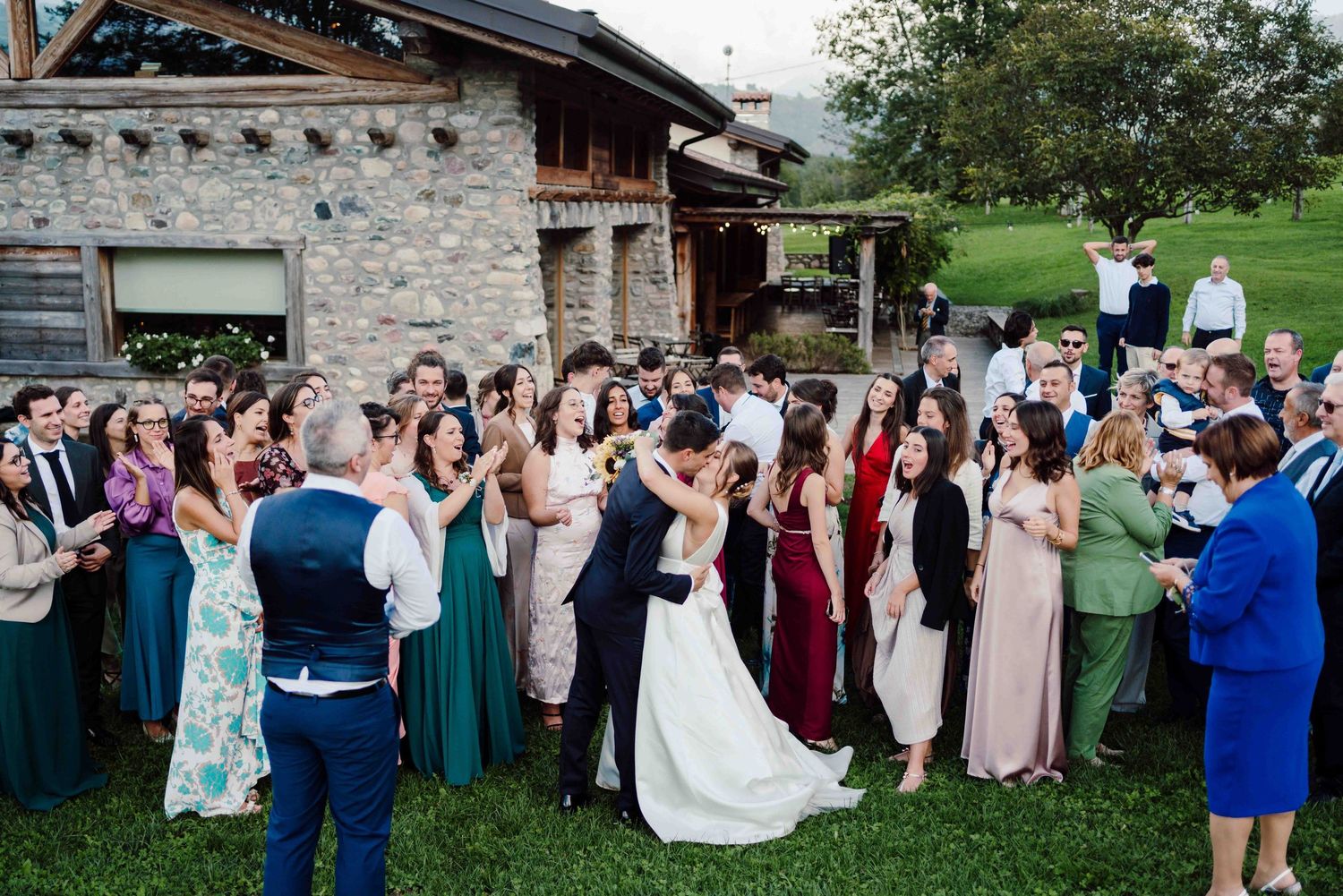 Wedding guests gather outdoors cheering as bride and groom share a kiss near a rustic stone venue.