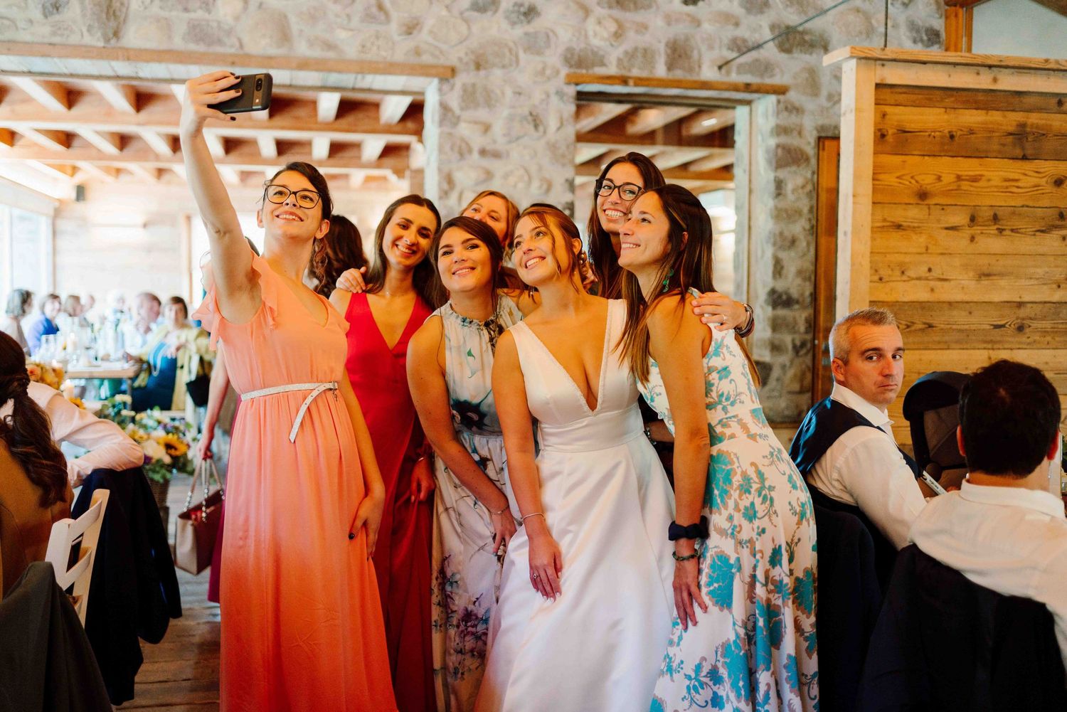 Bride in white dress takes selfie with bridesmaids in colorful gowns at rustic wedding reception venue.