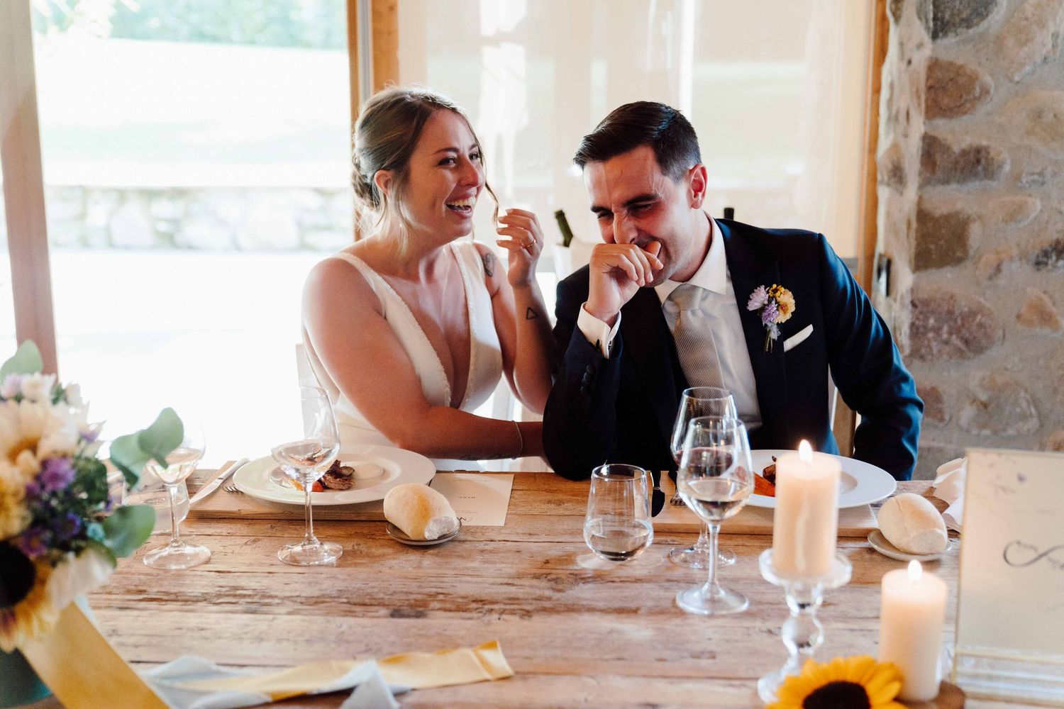 Bride and groom laughing together at rustic wooden wedding reception table with sunflowers and candles.