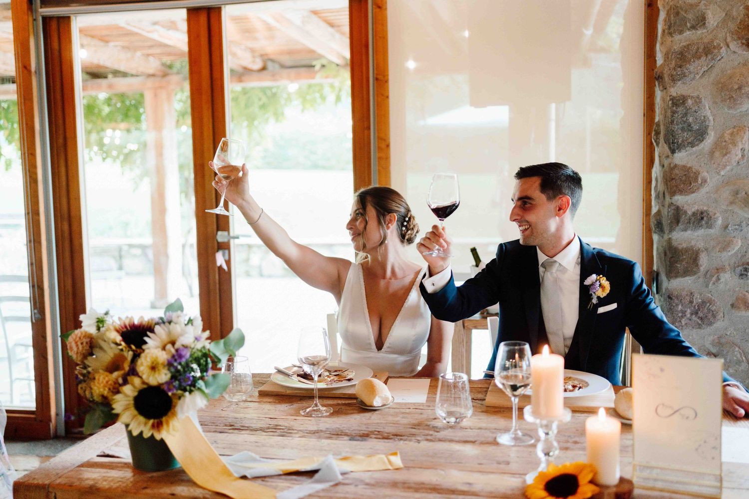 Bride and groom toasting with wine glasses at wedding reception table decorated with sunflowers and candles.