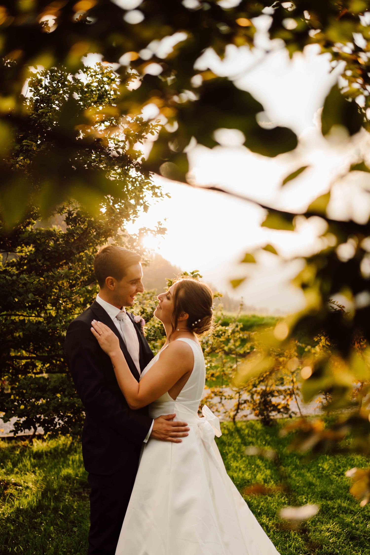 A bride and groom share a romantic moment under golden sunset light framed by lush green trees outdoors.