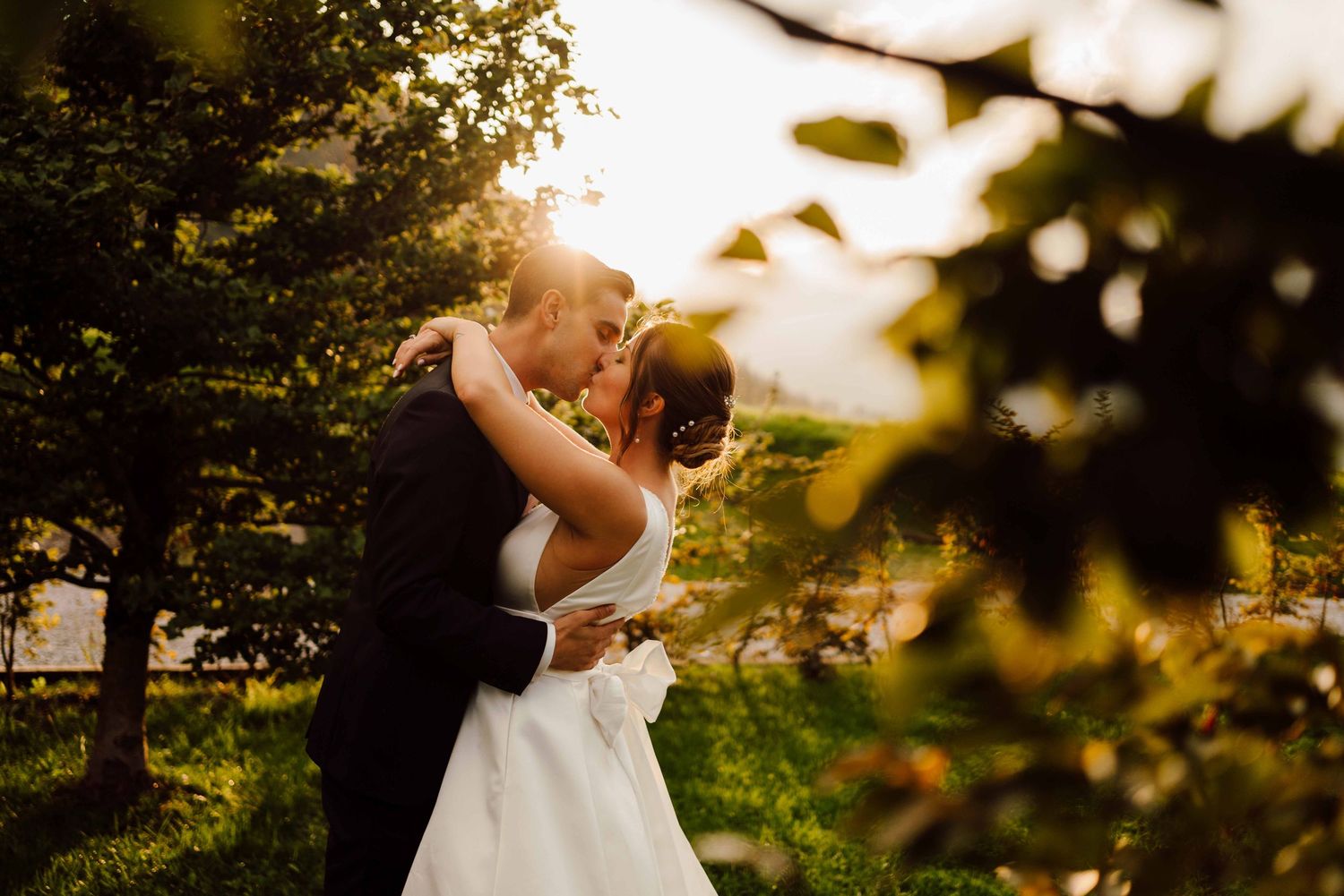 A bride and groom share a romantic kiss outdoors, surrounded by lush green trees with golden sunlight.