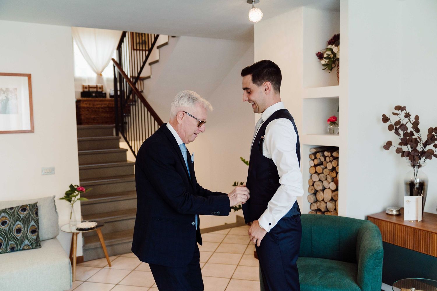 An older man helps a young groom adjust his suit in a bright home interior before a wedding ceremony.