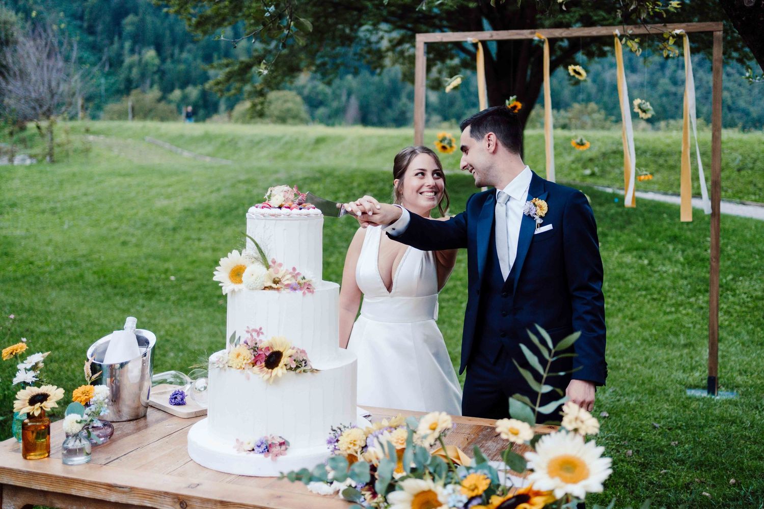 Bride and groom smiling while cutting a floral three-tier wedding cake outdoors with sunflowers and greenery.