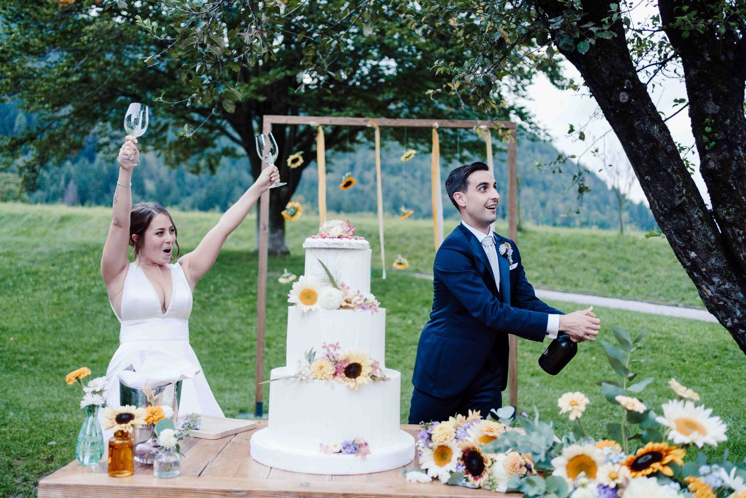 Joyful couple celebrating at wedding cake table with sunflowers, champagne, and a white tiered cake outdoors.