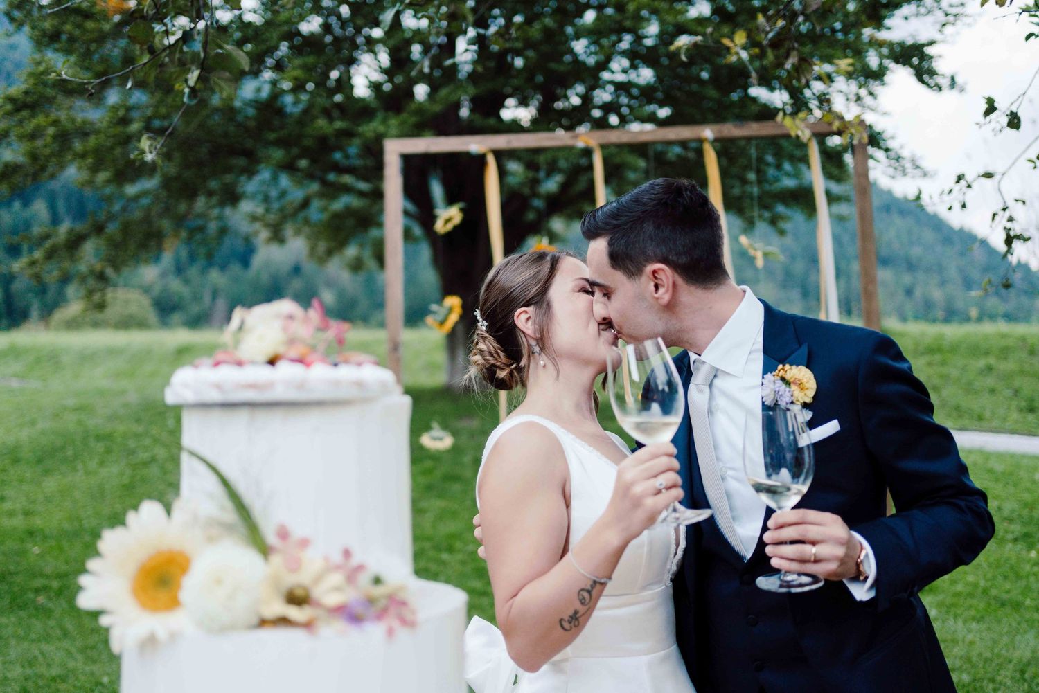 Newlywed couple kissing while holding champagne glasses beside a white wedding cake outdoors.