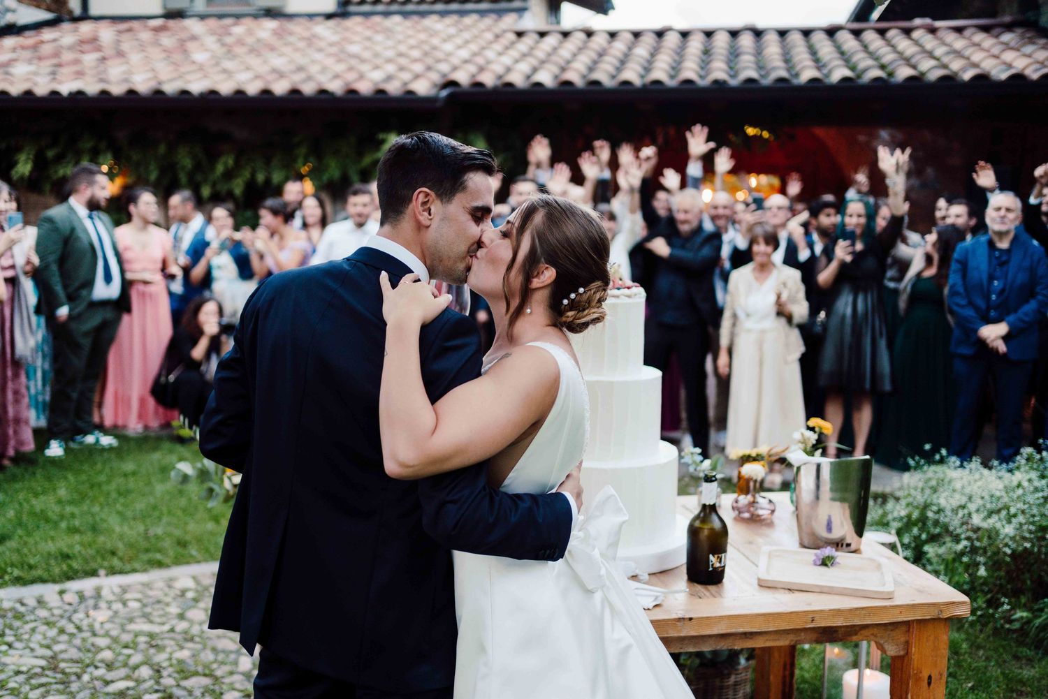 Bride and groom share first kiss at wedding reception with cheering guests and white tiered cake nearby.