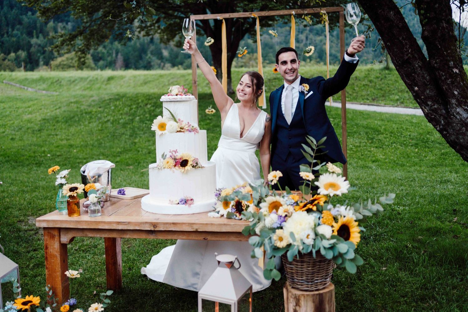 Bride and groom celebrate outdoors raising champagne glasses beside a floral wedding cake on a rustic wooden table.