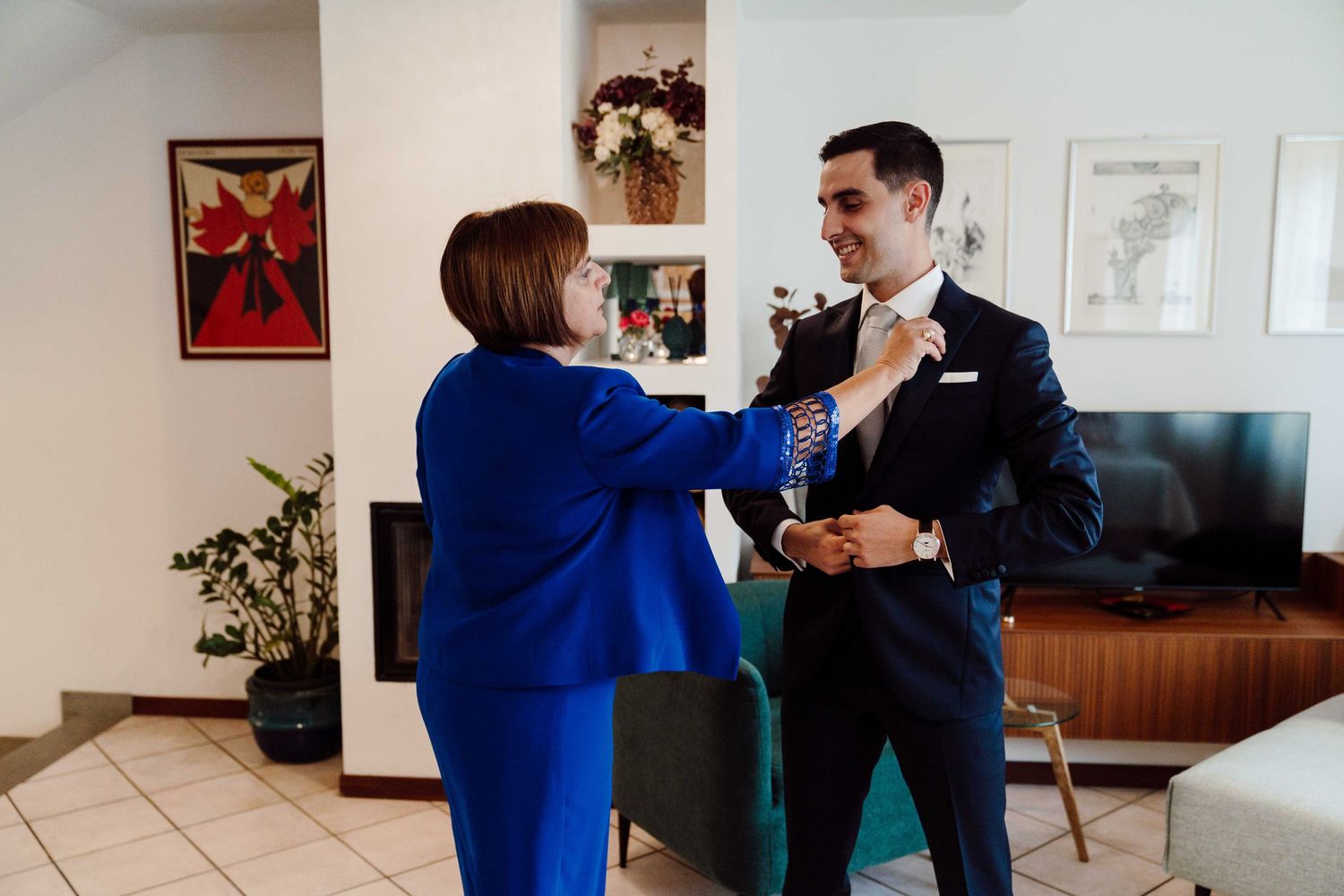 A woman in a blue suit adjusts a groom's tie as he prepares in a suit before his wedding ceremony.