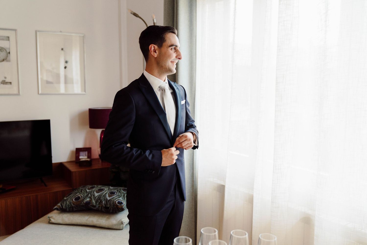 Groom in navy suit buttoning jacket by window with natural light in elegant hotel room.