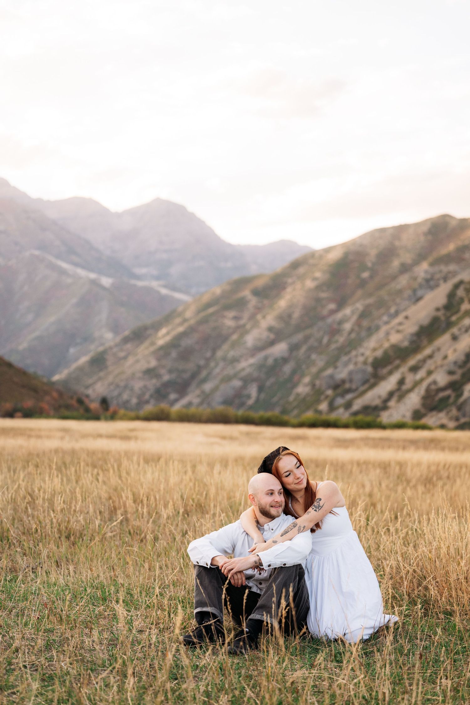 Couple sitting in golden field with mountain backdrop, woman in white dress embracing bald man at sunset.