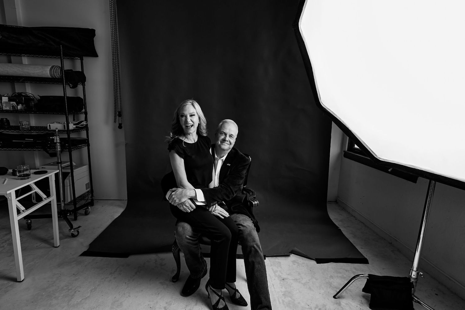 Black and white photo of a smiling couple sitting together in a professional photography studio with lighting equipment.