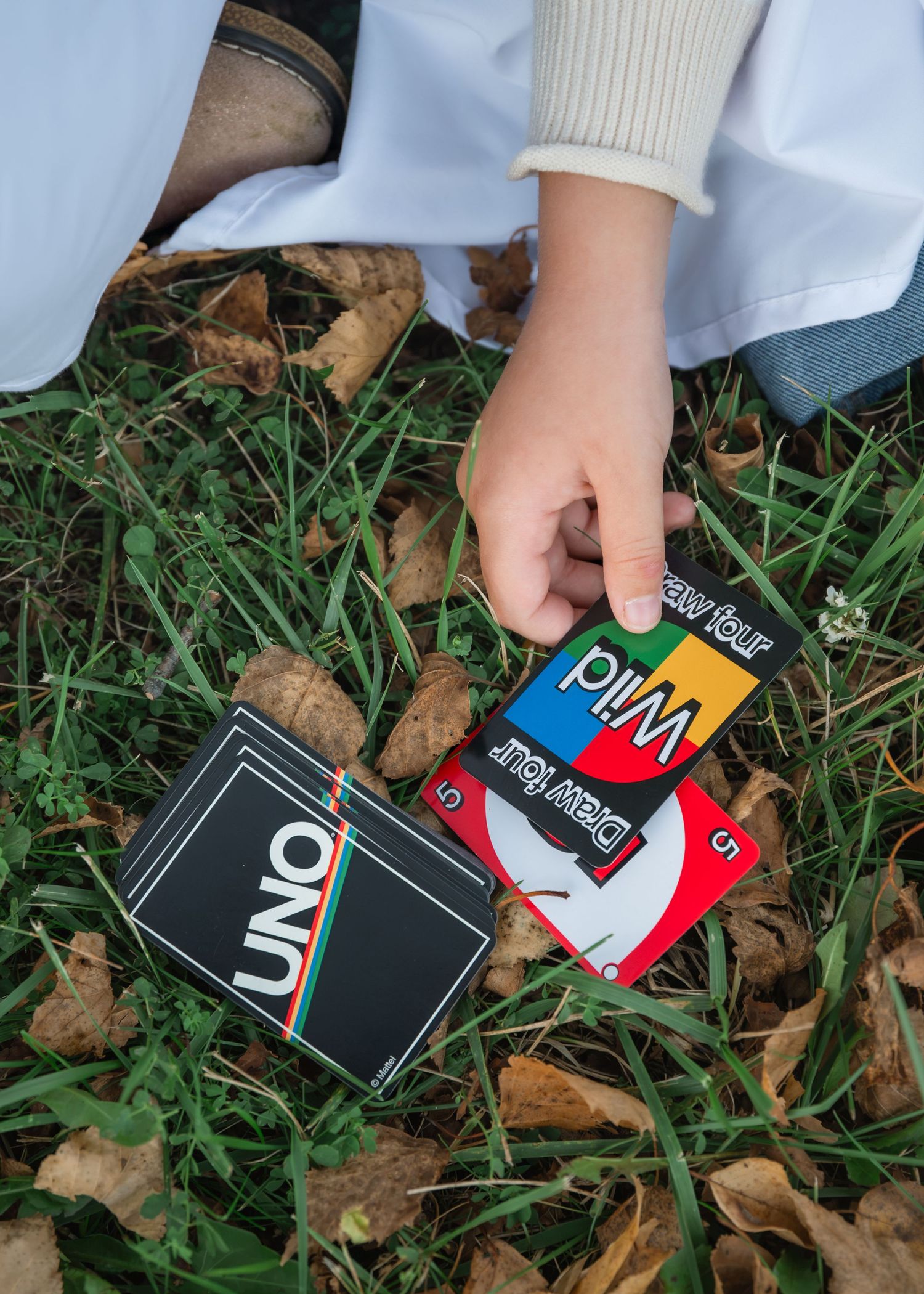 Child's hand placing a Wild Draw Four card beside an UNO deck on green grass scattered with fallen autumn leaves.