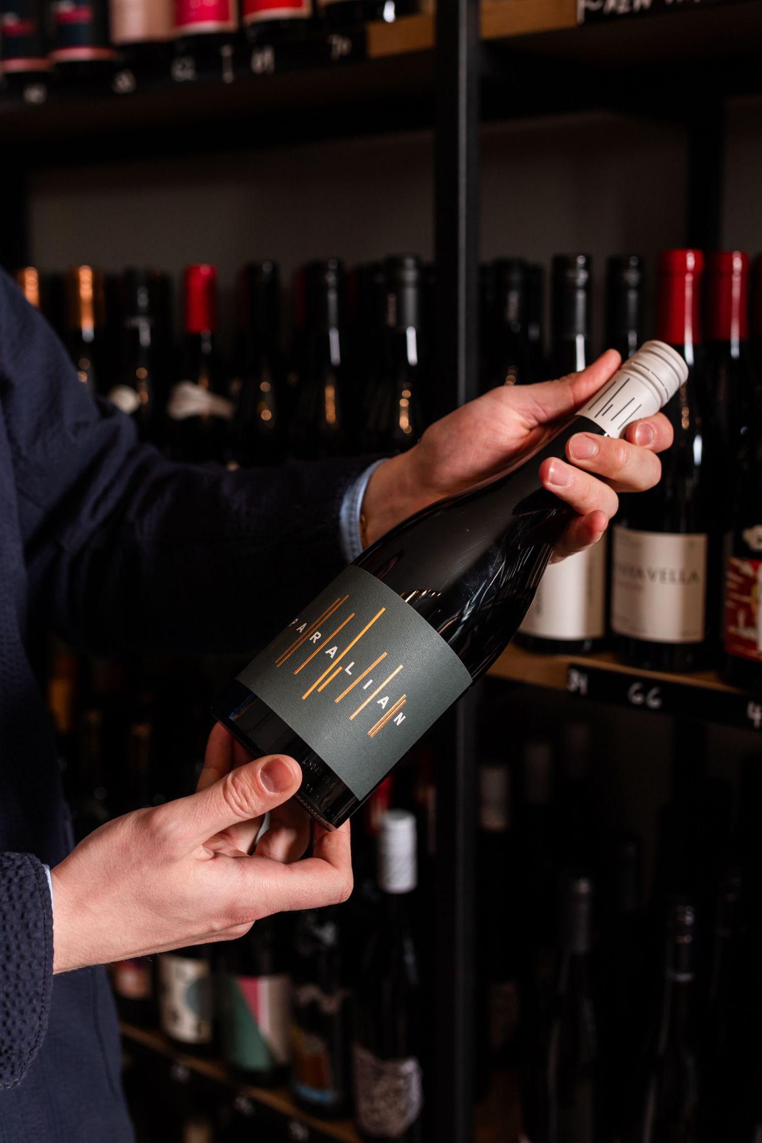 Person in a navy jacket holds a dark wine bottle with a gray label and gold line design in front of a shelf of wine bottles.