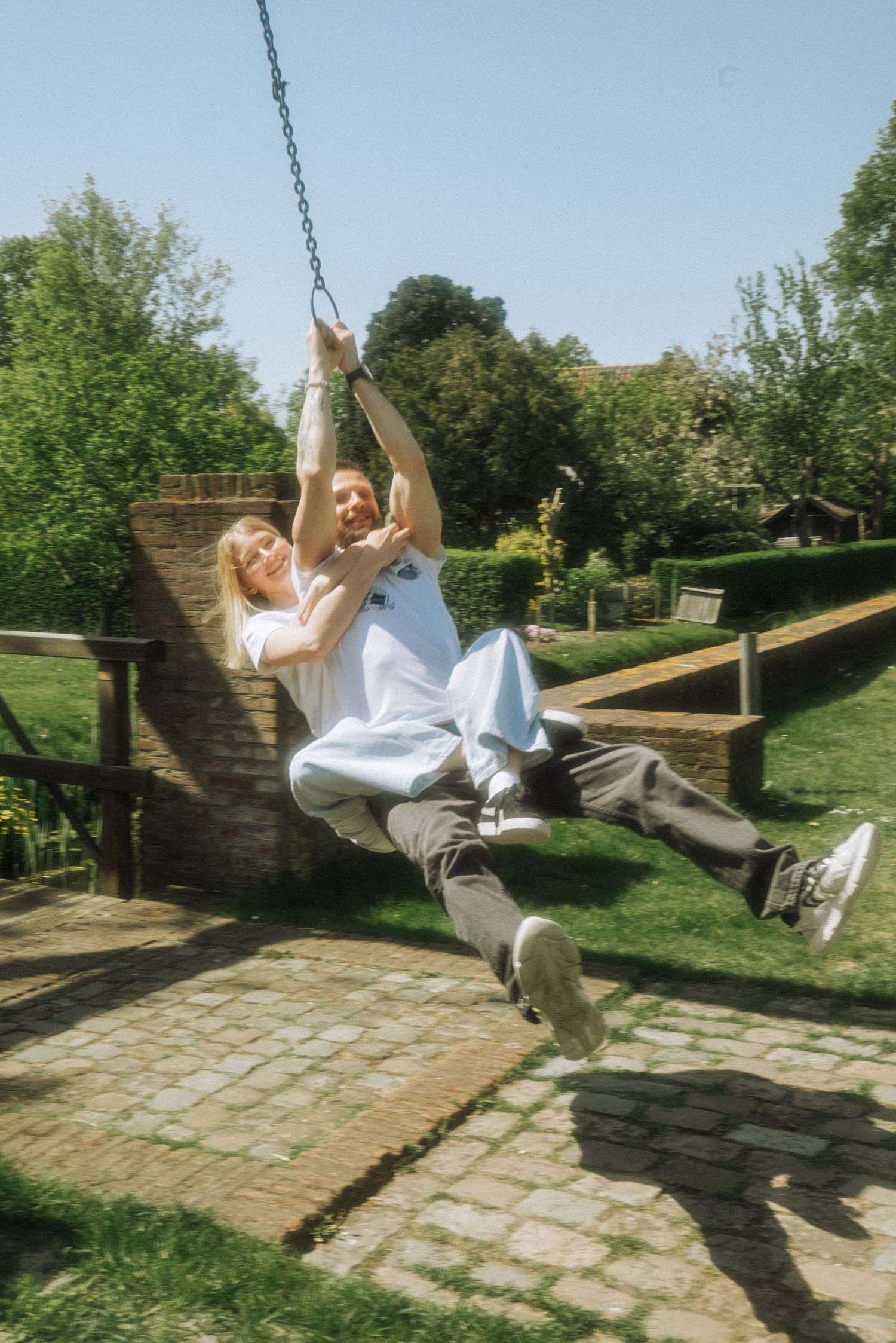 Two people laugh together while swinging on a chain-hung zip line in a sunny garden with brick paths, green trees. Huys Dever