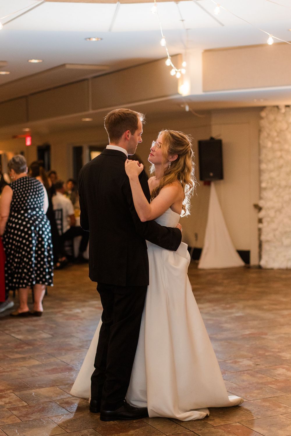 Bride and groom share their first dance at wedding reception, surrounded by guests in elegant ballroom with string lights.