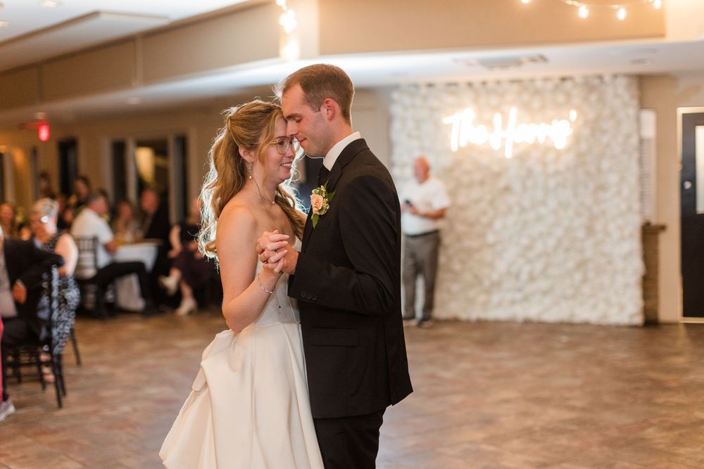Bride and groom share their first dance at elegant wedding reception with floral backdrop and guests seated nearby.