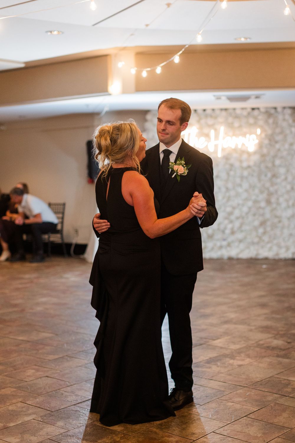 A groom in a black suit shares a mother-son dance with a woman in a black gown at a wedding reception.
