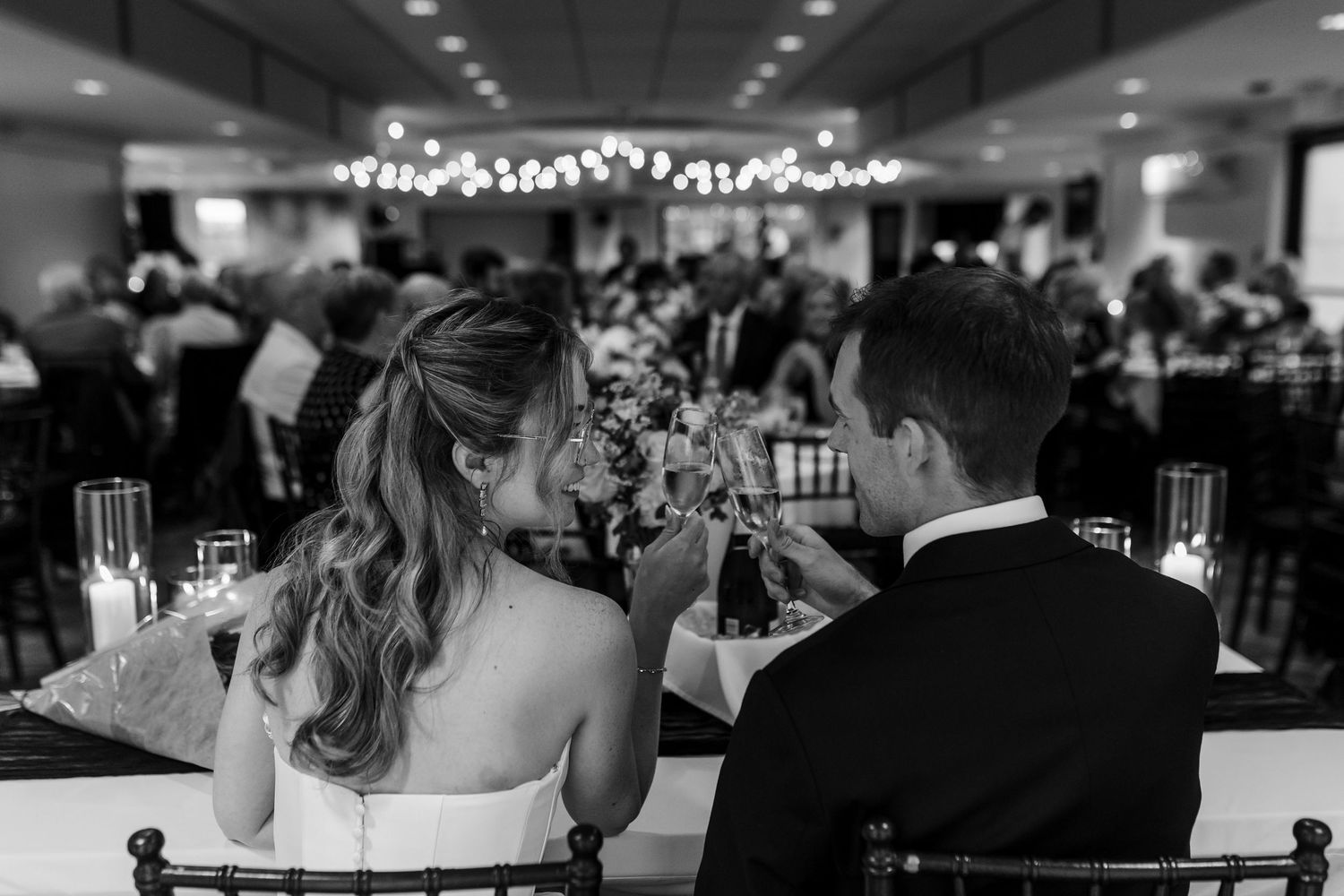 Black and white photo of bride and groom toasting at wedding reception with guests seated in elegant venue.
