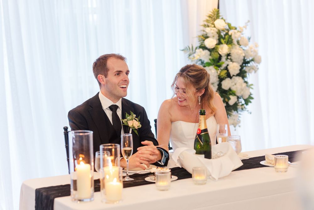 Bride and groom smiling at wedding reception table with champagne, candles, and white floral arrangements.