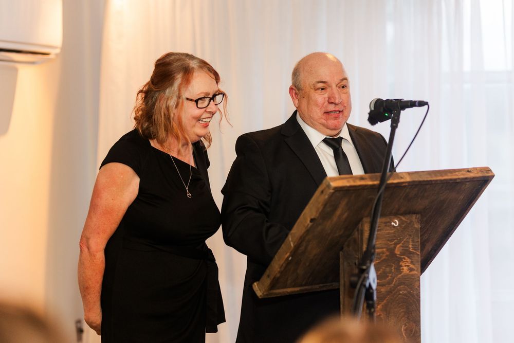 Couple in formal black attire speaking at a podium with microphone during an elegant event.