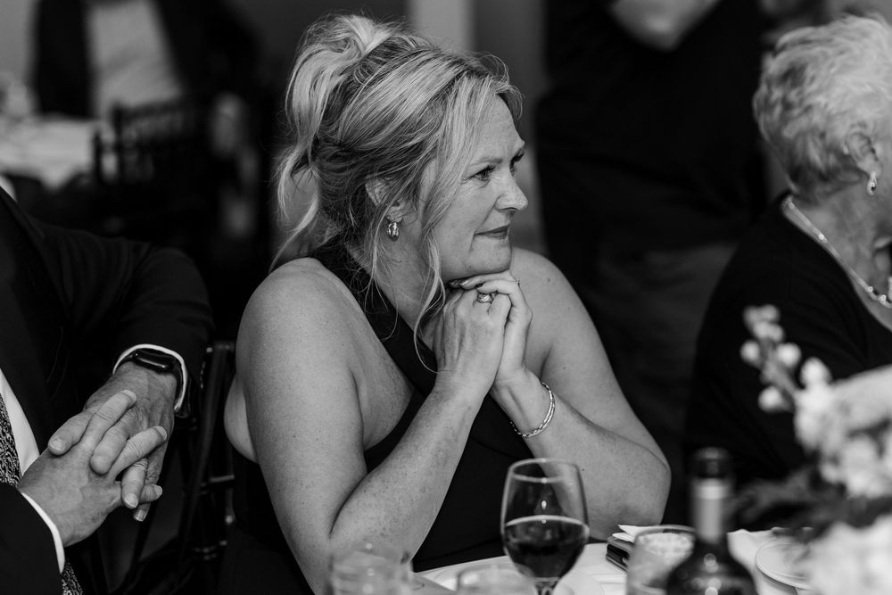 Black and white photo of a woman in a black dress sitting at a wedding reception table, hands clasped, looking thoughtfully ahead.