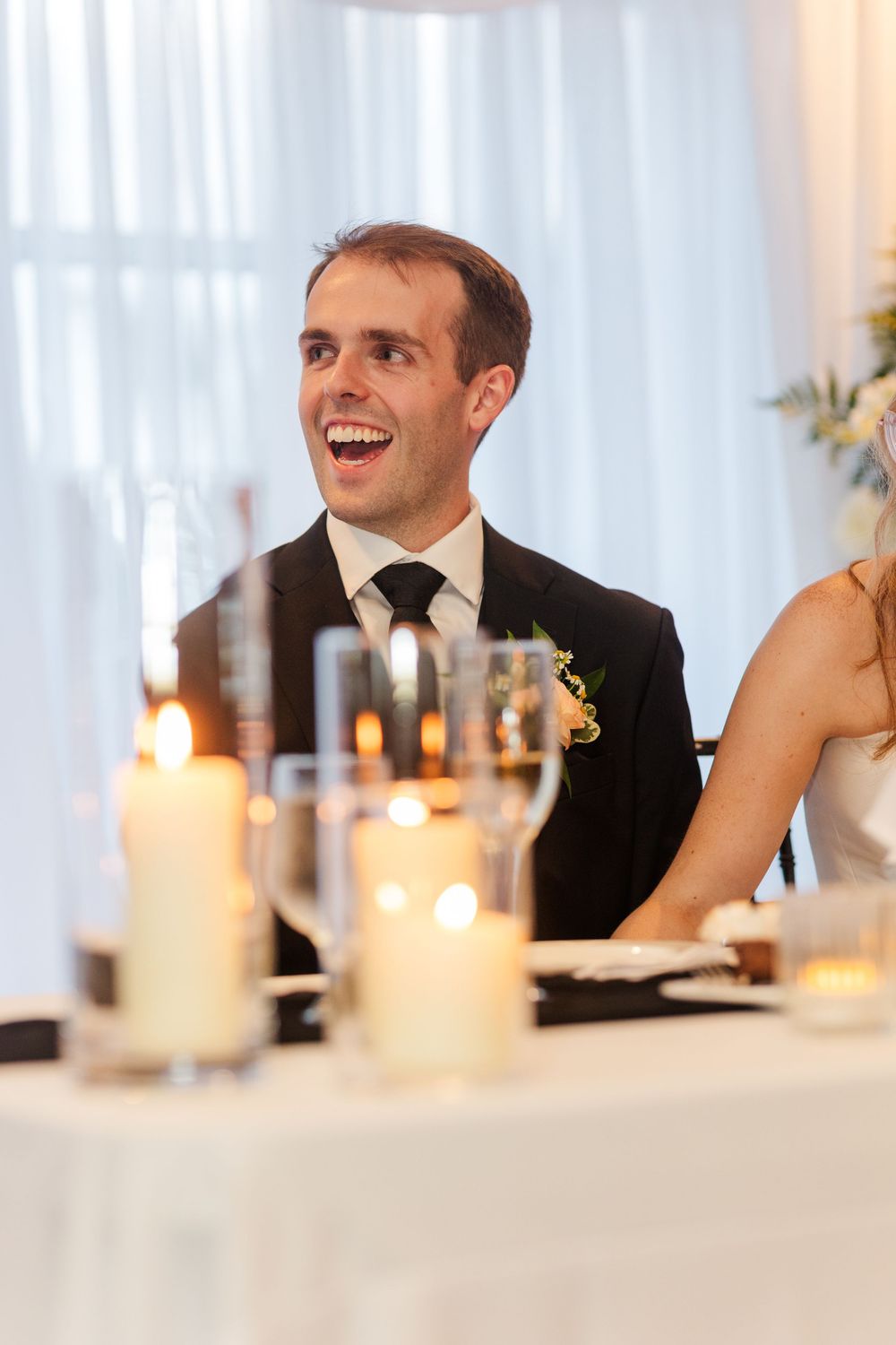Smiling groom in black suit with boutonniere seated at candlelit wedding reception table.