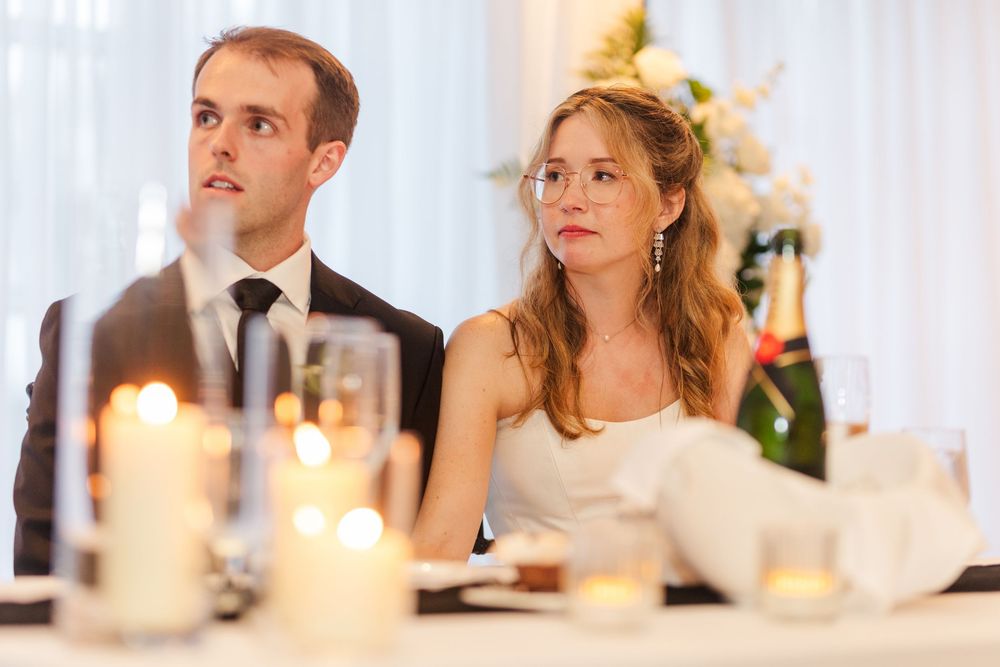 Bride and groom sitting at wedding reception table, looking attentively, surrounded by glowing candles.