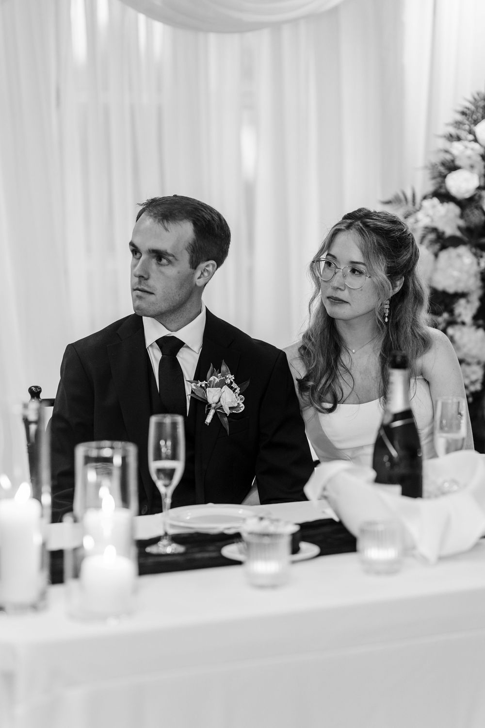 Black and white wedding photo of bride and groom seated at reception table with candles and champagne.