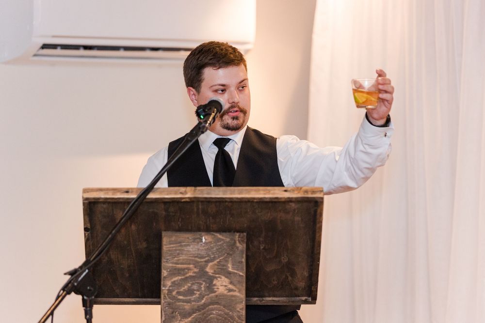 Man in vest raises glass of whiskey while giving a toast at a wedding reception podium with microphone.