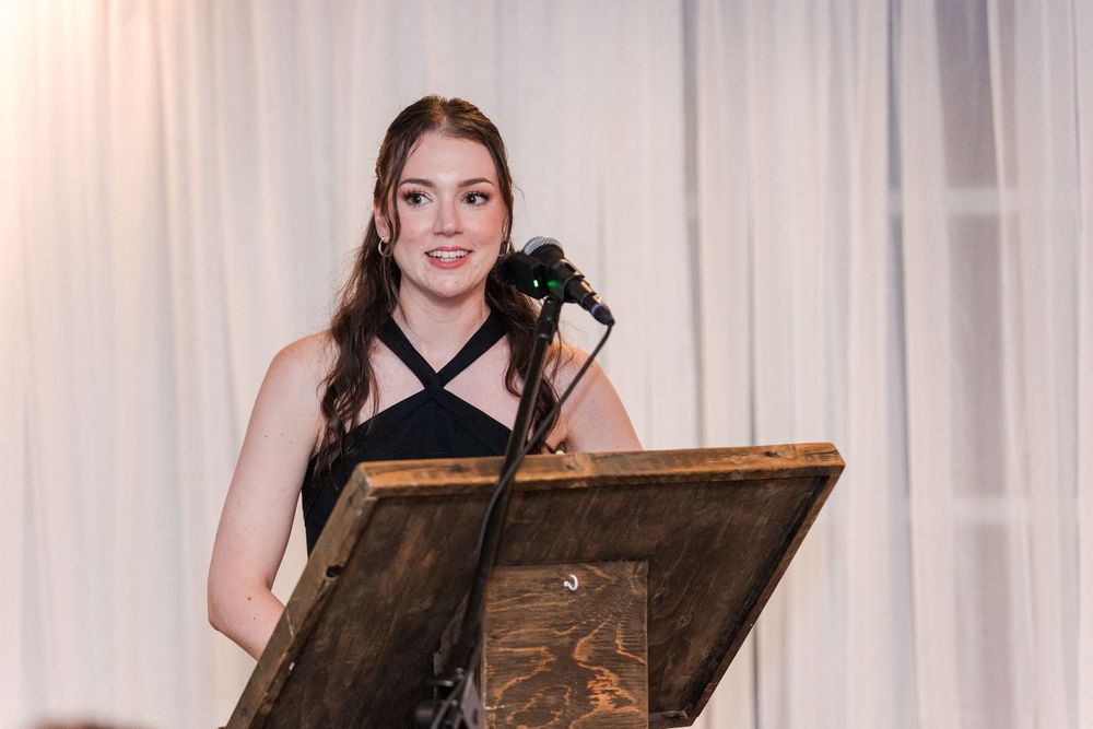 Young woman in black dress speaking at a rustic wooden podium with microphone at a formal event.